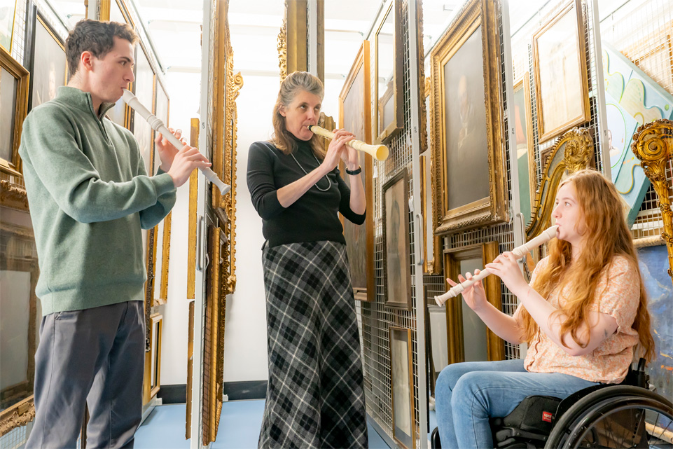 A female professor, wearing a black top and plaid skirt, playing a 3D printed clarinet, alongside a female student in a wheelchair and a male student, playing a 3D Printed Musical Instruments.